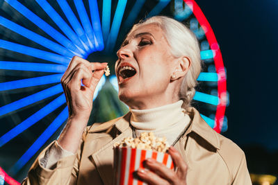 Smiling woman eating popcorn against ferris wheel at night