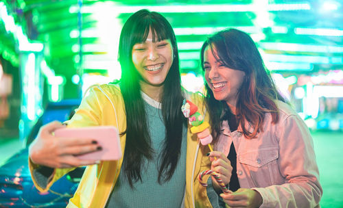 Cheerful woman taking selfie with friend while standing on illuminated street at night