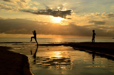 Silhouette boys at beach against cloudy sky during sunset