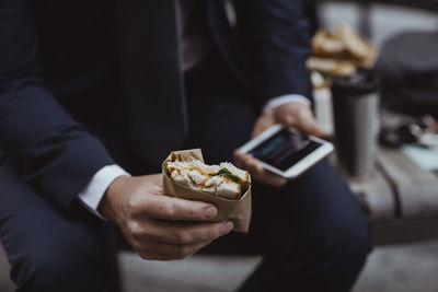 Midsection of entrepreneur with smart phone and sandwich sitting in city