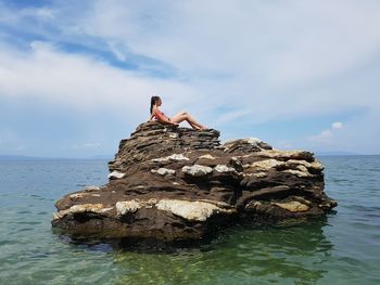 Side view of girl lying on rock in sea against cloudy sky