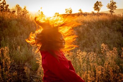 Side view of woman tossing hair while standing by plants against sky during sunset