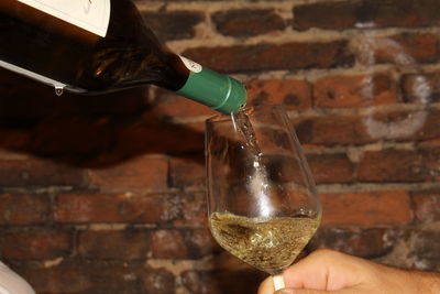 Close-up of hand pouring wine in glass against brick wall