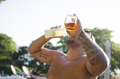 Midsection of shirtless man drinking glass against sky