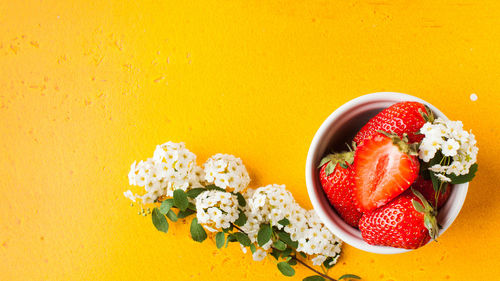 Ripe strawberries whole and cut in a white bowl on a yellow background surrounded by white flowers.