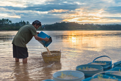 Side view of man pouring water in basket in lake during sunset