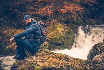 Side view of mid adult man with backpack sitting on mountain by waterfall