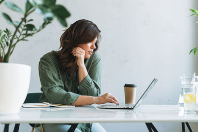 Young thinking brunette woman working at laptop on table with plant in the bright modern office