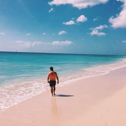 Full length rear view of shirtless man walking at beach against sky on sunny day