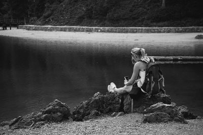 Rear view of woman photographing from mobile phone while sitting on rocks at lakeshore