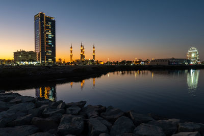 Illuminated buildings by river against sky during sunset