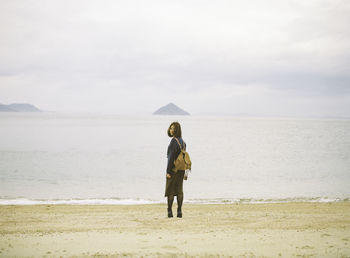 Full length of young woman standing at beach against cloudy sky