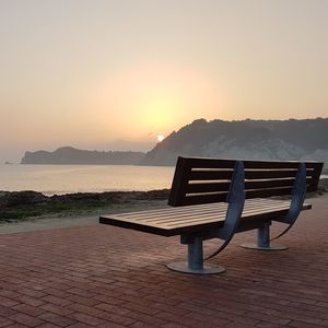 Empty bench on beach against sky during sunset