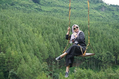 Full length portrait of woman sitting on rope swing in forest