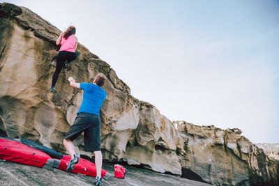 Man looking at woman climbing on rock at beach against sky