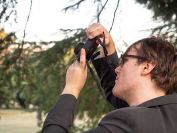 Portrait of young man holding camera against trees