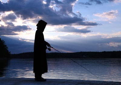 Silhouette man fishing by lake against sky during sunset