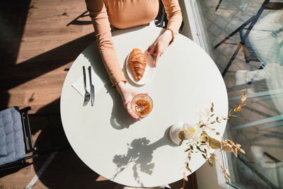 Anonymous female in beret sitting at table in cafe with aromatic glass of coffee and freshly baked croissant