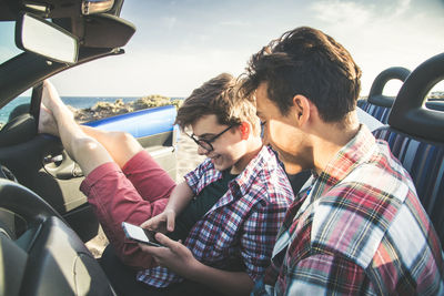 Teenage boys using mobile phone in car against sky
