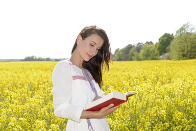 Beautiful woman reading book while standing by oilseed rape field