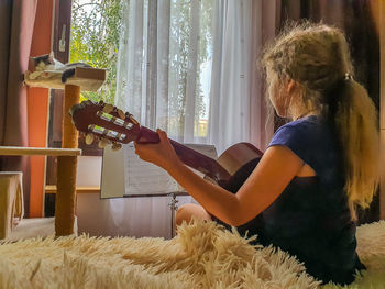 Girl playing guitar while sitting on bed at home