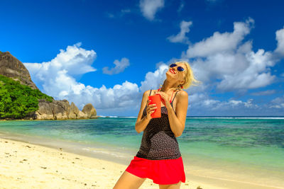 Happy woman holding drink at beach against sky