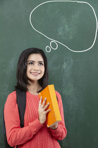 Thoughtful female student holding book by speech bubble on blackboard