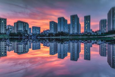 Modern buildings reflecting in river against sky during sunset