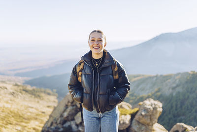 Smiling young woman standing on mountain against sky during sunny day