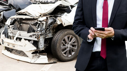 Midsection of businessman using mobile phone while standing against damaged car