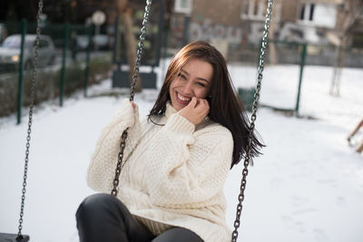 Portrait of happy young woman swinging in snow covered playground