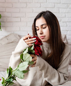 Young caucasian brunette woman sitting in the bed celebrating valentine day smelling red roses