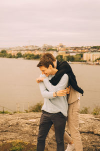 Cheerful male friends embracing while standing on rock formation against lake