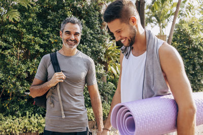 Portrait of smiling man standing with male friend holding yoga mat at wellness resort