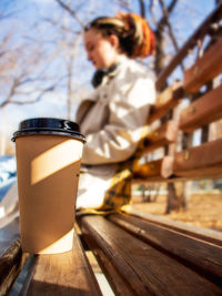 Coffee in a cup stands on a bench in the park and a young modern woman in a raincoat 