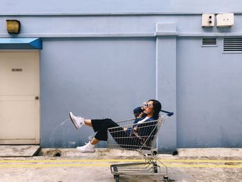 Side view of young woman sitting in shopping cart against wall