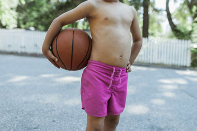 Midsection of shirtless boy holding basketball while standing on footpath at park