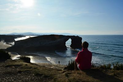 Rear view of man sitting on cliff by sea against sky