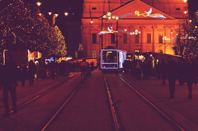 Illuminated tramway moving on street amidst people against decorated buildings