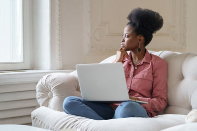 Woman looking away while sitting with laptop on sofa at home