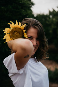 Side view of brown haired woman in light clothing looking at camera holding sunflower behind ear amid green trees during summer vacation
