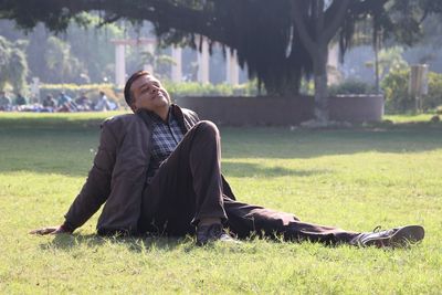 Mid adult man sitting on grassy field at park during sunny day