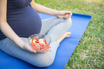 Low section of pregnant woman holding bowl with fruits while sitting on exercise mat