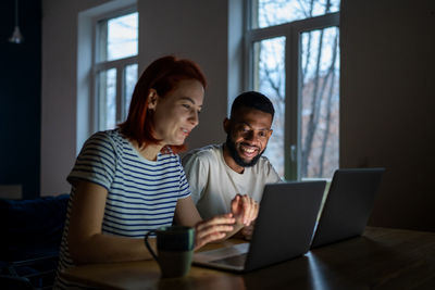 Smiling multiracial family couple enjoy freelancing, sitting at table working on laptops together