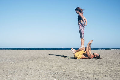 Couple practicing yoga at beach against clear blue sky