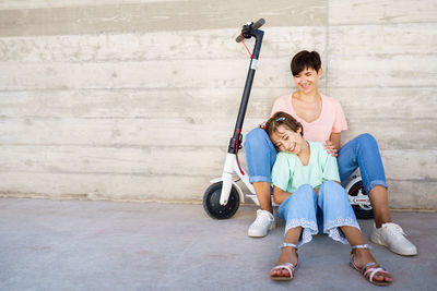 Mother with daughter sitting on push scooter against wall