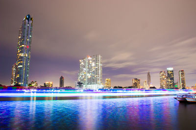 Illuminated modern buildings by river against sky at night