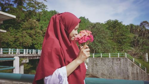 Woman holding pink flower against trees and plants