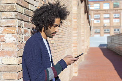 Young man using phone while standing against brick wall