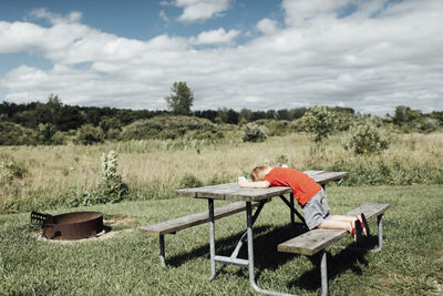 Side view of boy lying on picnic bench against cloudy sky during sunny day
